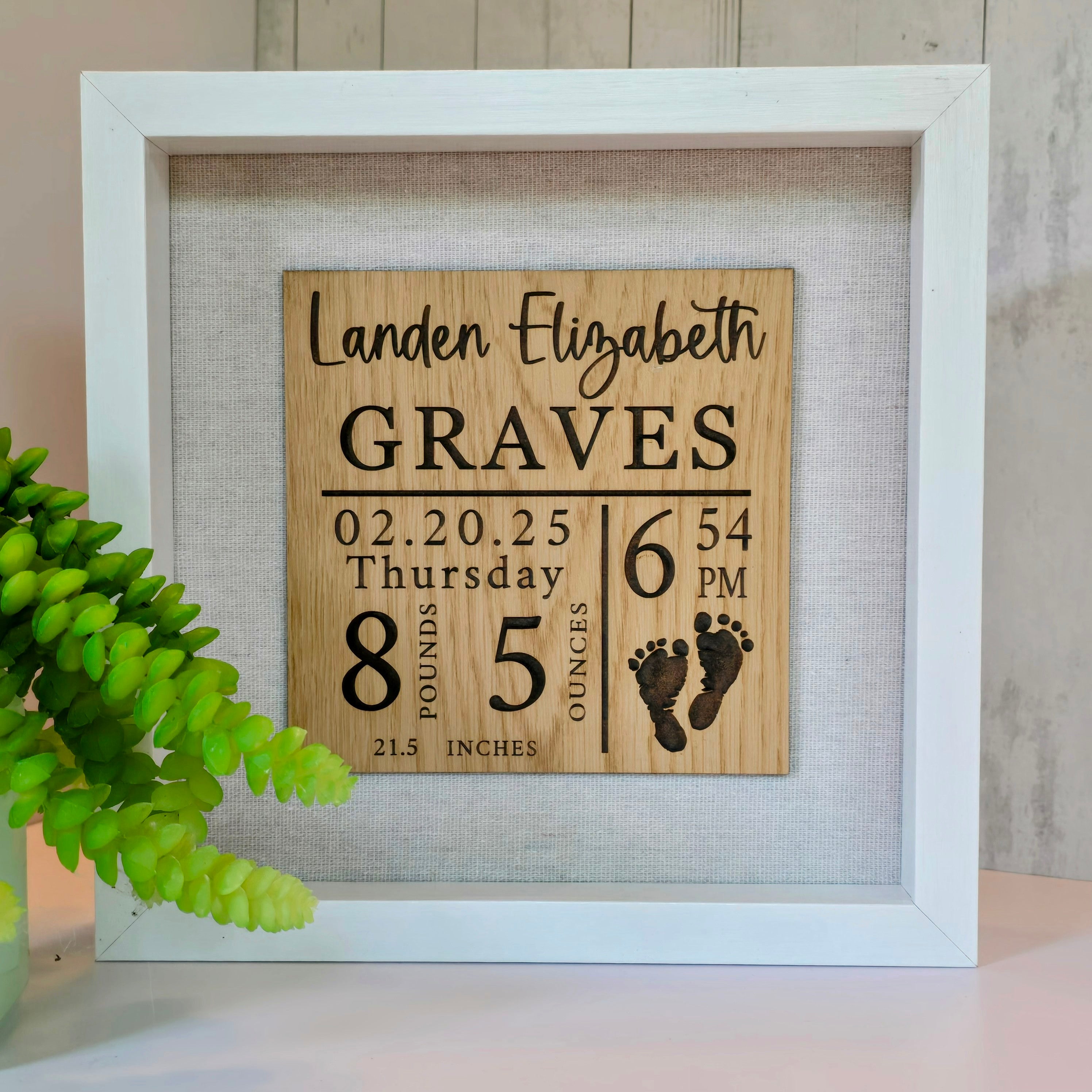 A personalized birth announcement shadow box with the name, birth date, weight, and length of a baby. The box is framed and displays this information on a wooden panel with a white background, accompanied by a silhouette of a foot.