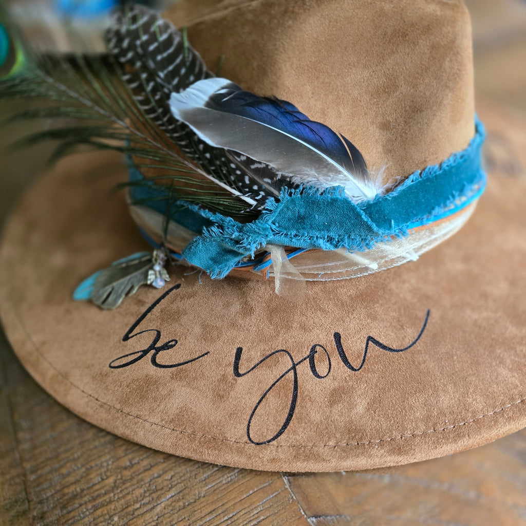 A tan suede cowboy-style hat with a blue and turquoise feathered band, blue feather accents, and the words 'be you' embroidered in black script on the brim, shown on a wooden surface. Hat Burning and Hat Bar Workshop by Rise and Shine Gifts in Dothan, Alabama.