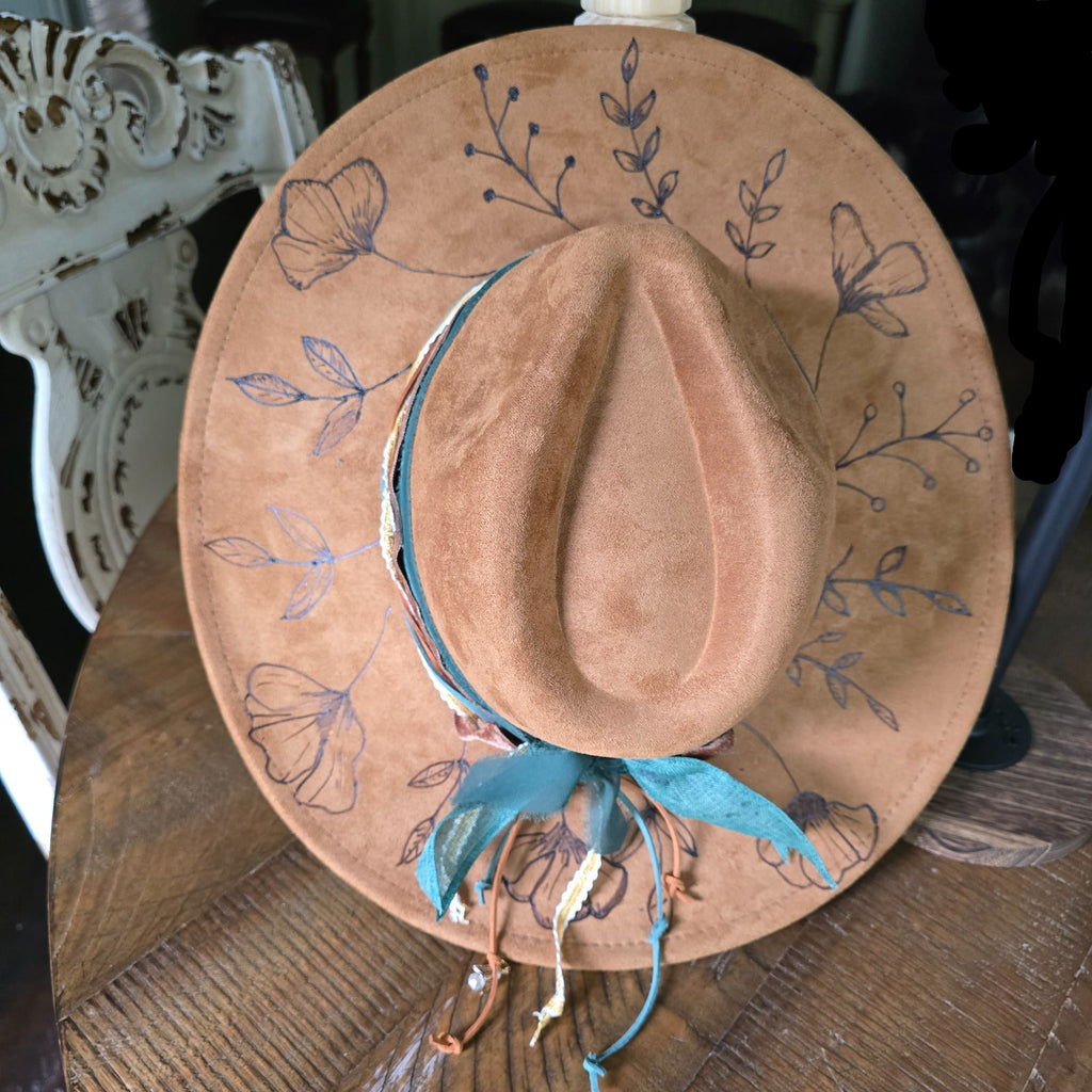 A tan suede cowboy hat with hand-painted floral designs and a turquoise ribbon. Hat Burning and Hat Bar Workshop by Rise and Shine Gifts in Dothan, Alabama.