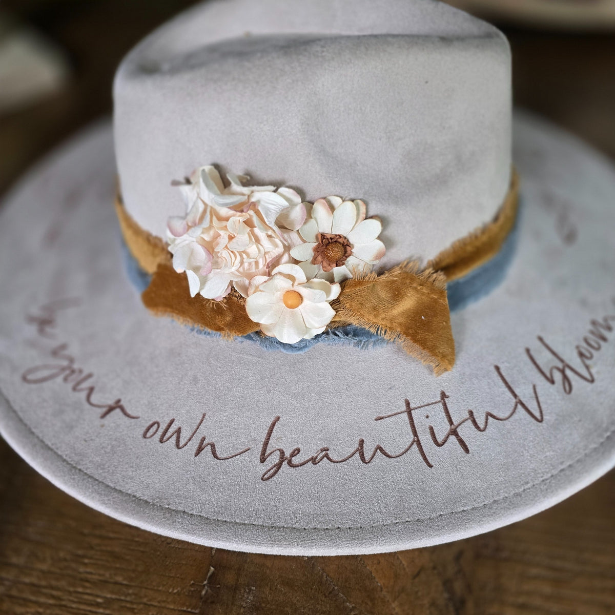 A light gray felt hat with a wide brim, decorated with a tan ribbon, blue fabric trim, and white and peach fabric flowers. The brim features brown cursive text reading 'be your own beautiful bloom'.  Hat Burning and Hat Bar Workshop by Rise and Shine Gifts in Dothan, Alabama.
