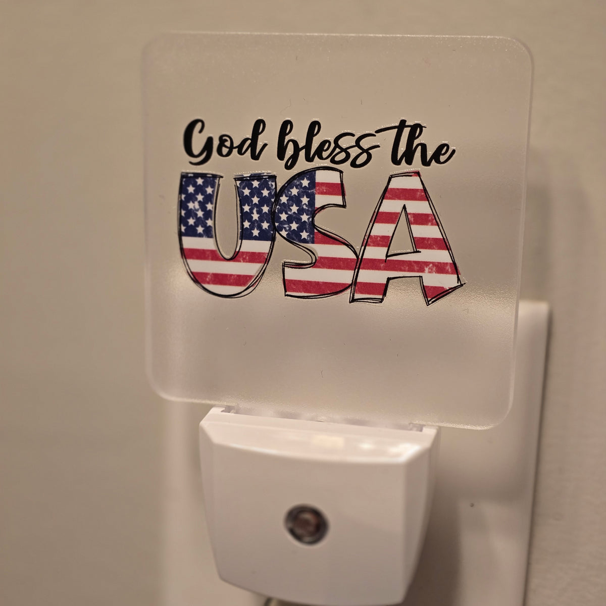 Frosted acrylic night light with a "God Bless the USA" American flag design, plugged into a wall outlet