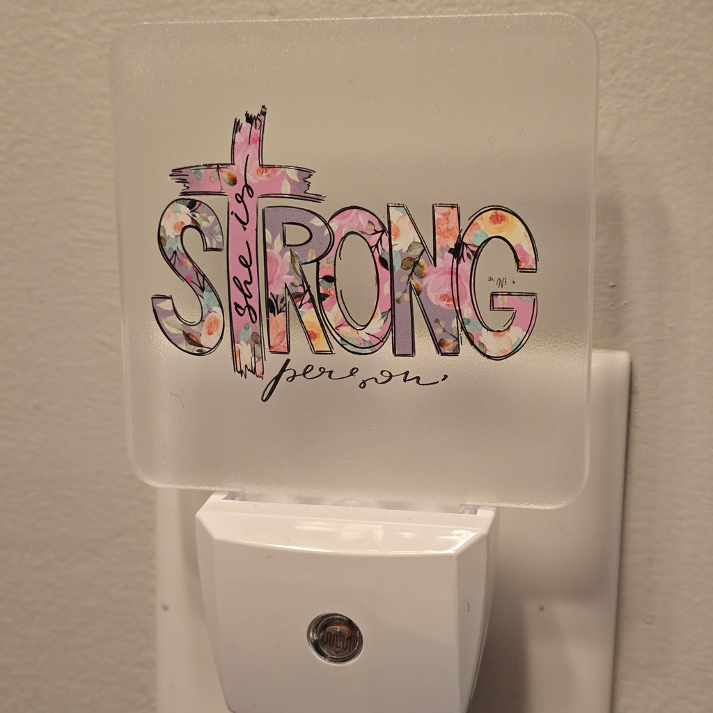 Frosted acrylic night light with a pink floral "She is Strong" cross design, plugged into a wall outlet