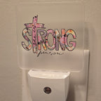 Frosted acrylic night light with a pink floral "She is Strong" cross design, plugged into a wall outlet