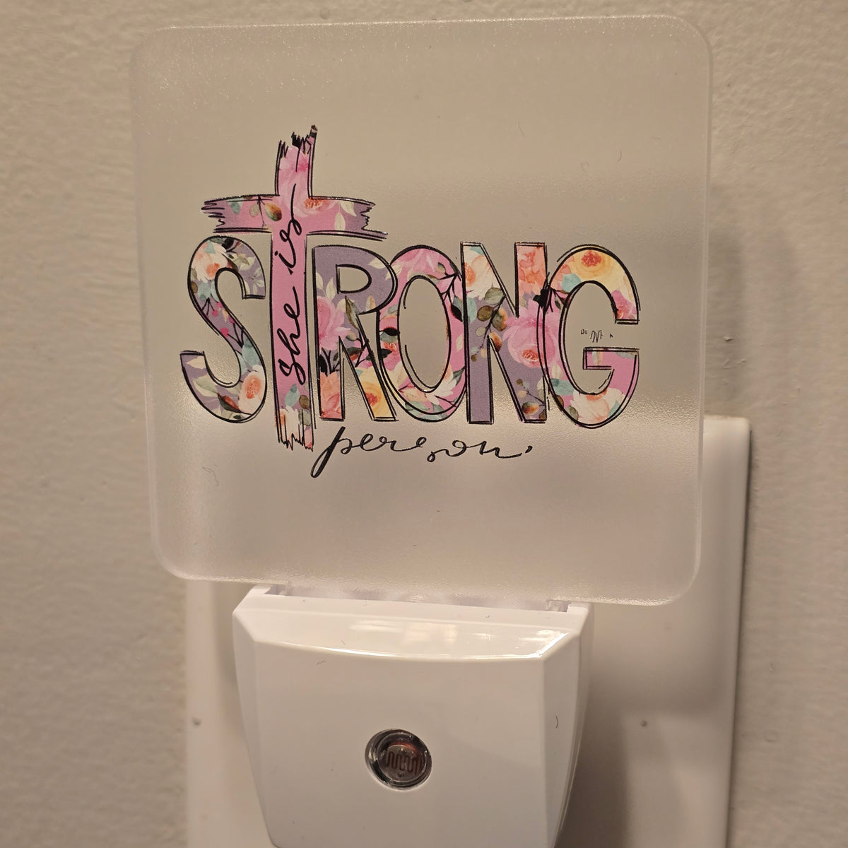 Frosted acrylic night light with a pink floral "She is Strong" cross design, plugged into a wall outlet