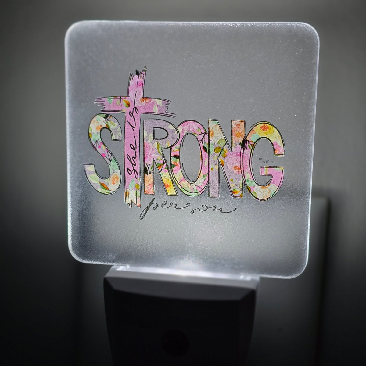 Frosted acrylic night light with a pink floral "She is Strong" cross design, glowing in a dark room