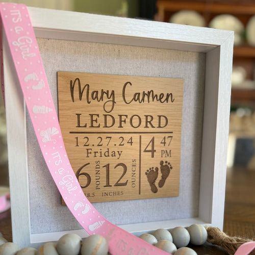 A personalized birth announcement shadow box with the name, birth date, weight, and length of a baby. The box is framed and displays this information on a wooden panel with a white background, accompanied by a silhouette of a foot.