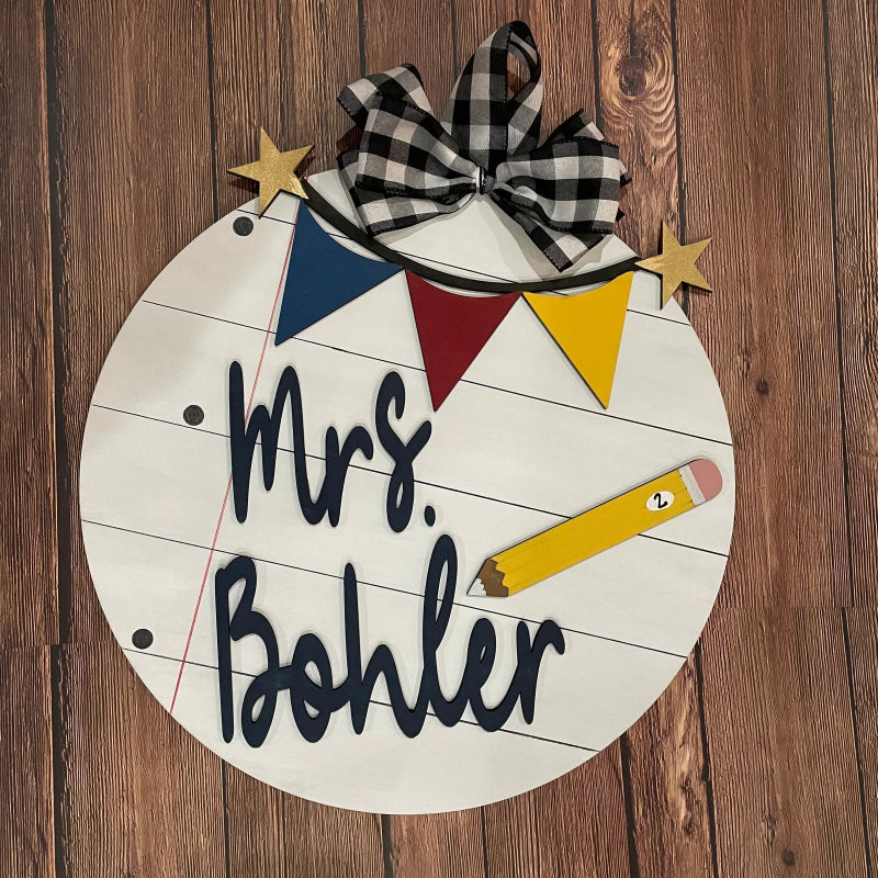 Round painted 3D wooden door hanger with 'Mrs. Bohler' text, colorful flags, and a 3D pencil. Paint this design at a Paint Party in Dothan, Alabama or order a DIY kit.  Paint the flags in your school colors!