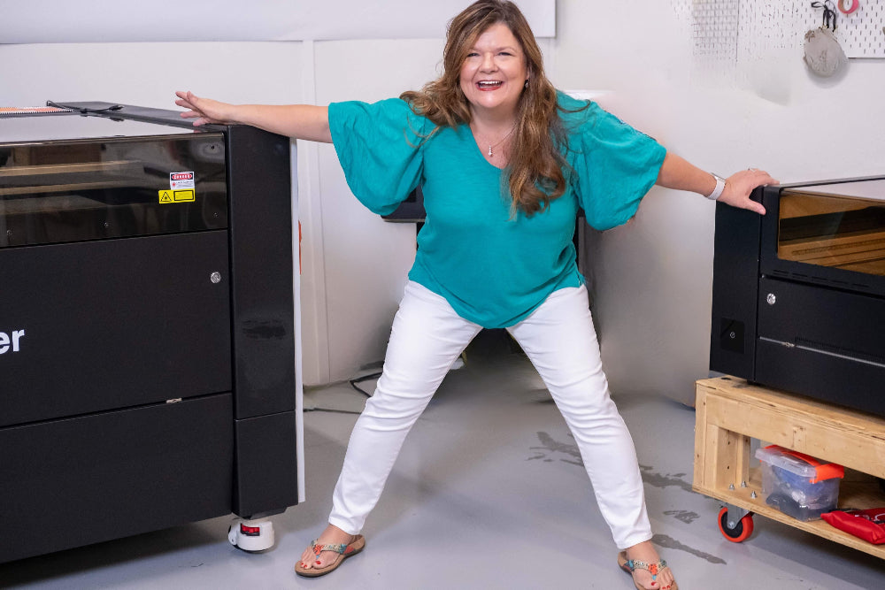 Regina attending a Laser Course, in teal shirt and white pants posing in front of large laser machines. Rise and Shine Gifts