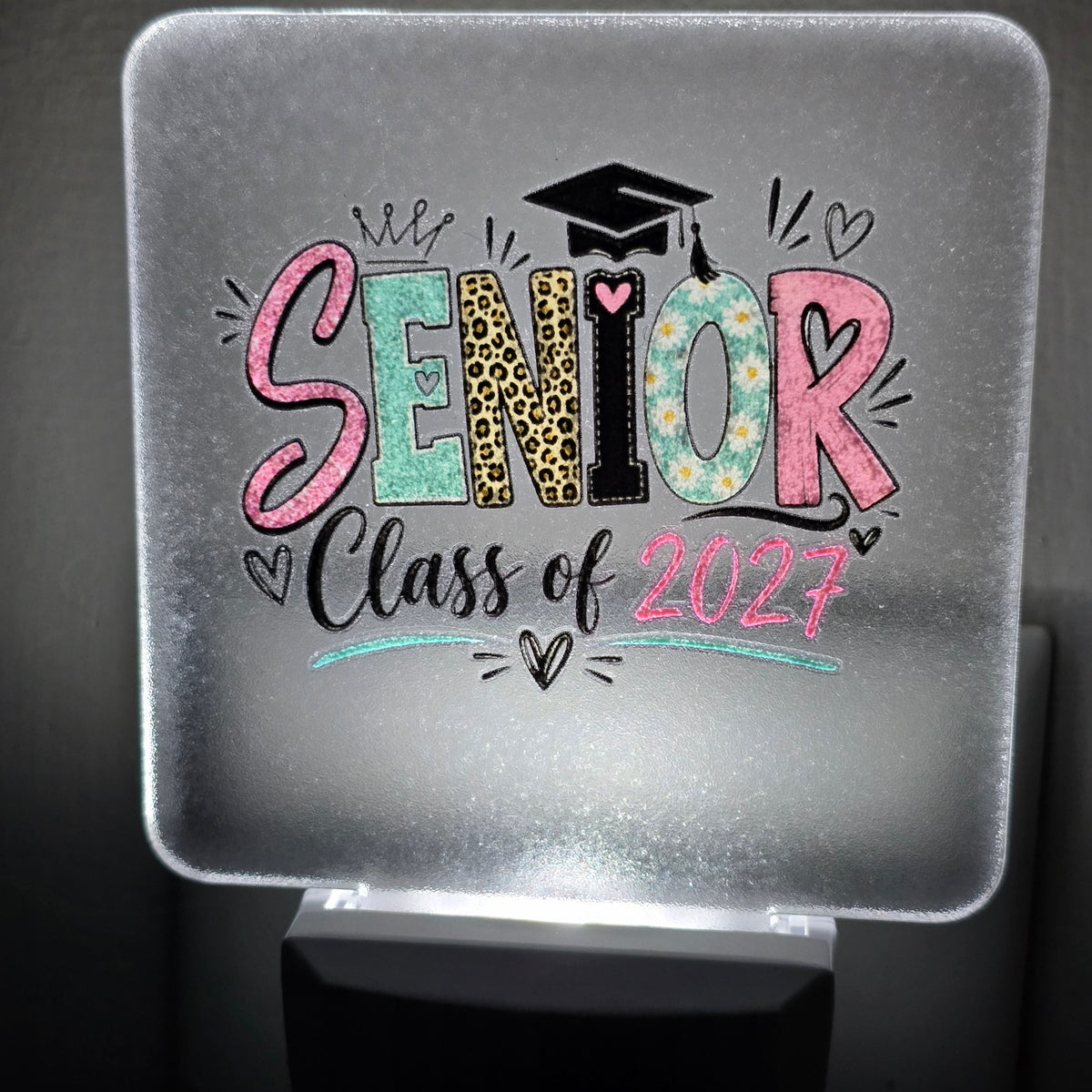 A square frosted glass night light featuring a colorful graduation-themed graphic that reads 'SENIOR Class of 2027' with a black graduation cap icon, shown glowing against a dark background. UV-printed by Rise and Shine Gifts in Dothan, Alabama.